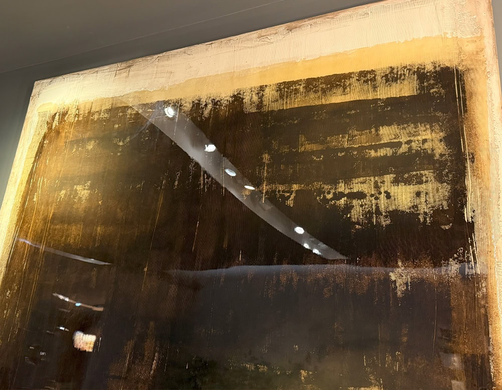 The Dra Sintensi Art is presented in a close-up view, highlighting textured brushstrokes in deep browns and golds. Reflections on the glossy surface add depth and dimension to the abstract composition.